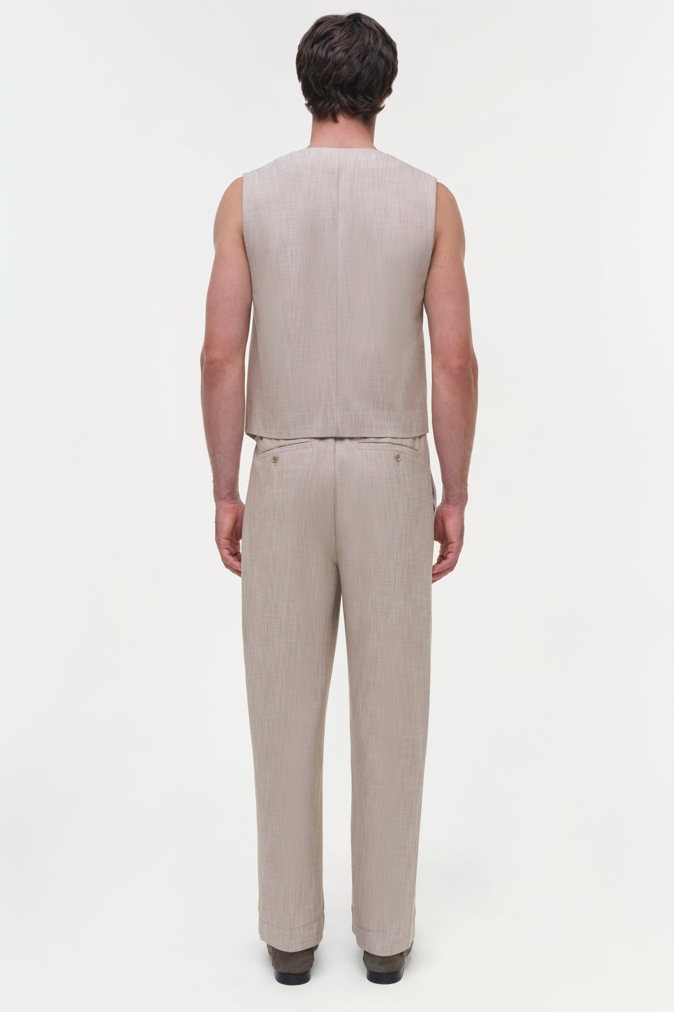Man wearing beige sleeveless vest and matching trousers, shown from behind against white background.