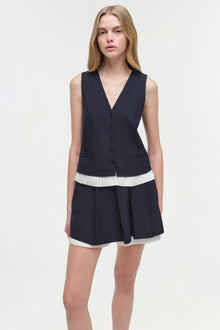 Woman wearing navy sleeveless button-front vest with white pleated trim over navy pleated skort with white hem.