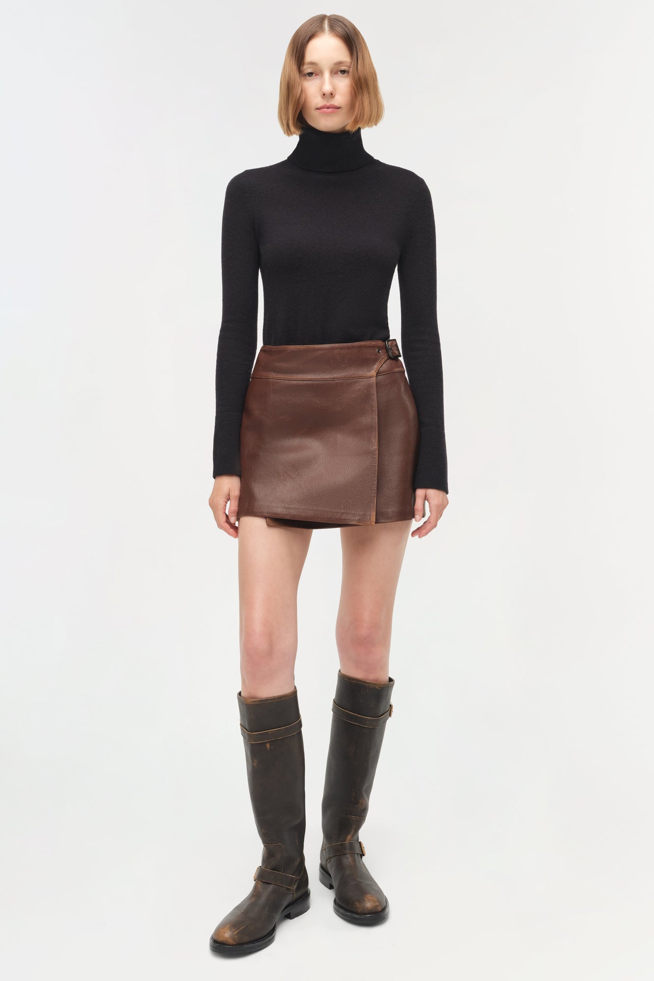 Brown leather mini skirt with wrap-front design and buckle closure, paired with black turtleneck and boots.