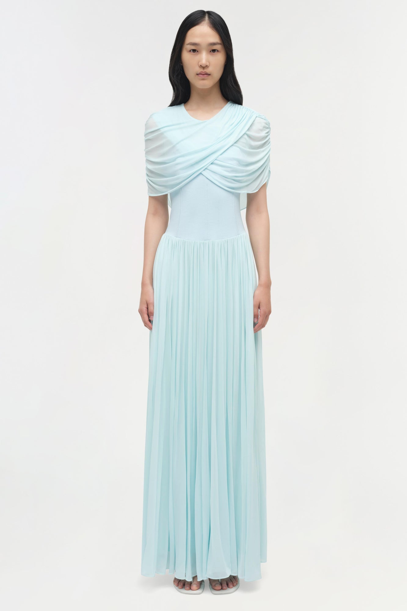 Light blue jersey maxi dress with draped cape-style overlay across shoulders and pleated skirt.