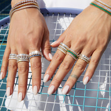 Lifestyle Model Close-up of a woman's hands resting on the light blue strings of a tennis racket, wearing multiple stacked gold rings and bracelets featuring diamonds, green stones, and blue stones