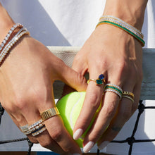 Lifestyle Model Close-up of a woman's hands holding a tennis ball against a tennis net, wearing multiple stacked gold rings and bracelets featuring diamonds, green stones, and blue stones.