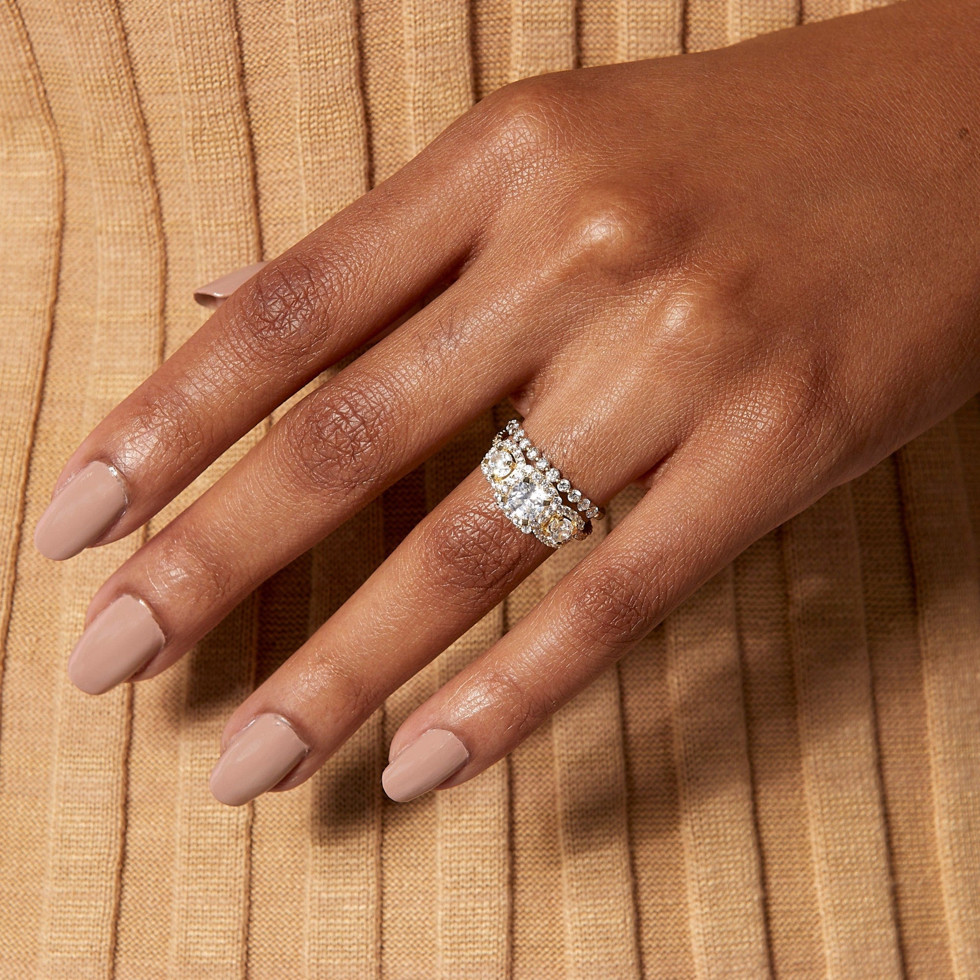 Lifestyle Model Close-up of a woman's hand resting gracefully against a ribbed, golden-beige fabric. On her ring finger, she is wearing a striking stack of three rings: a prominent cushion-cut halo diamond ring nestled between two diamond bands. Her nails are painted in a soft, neutral pink.
