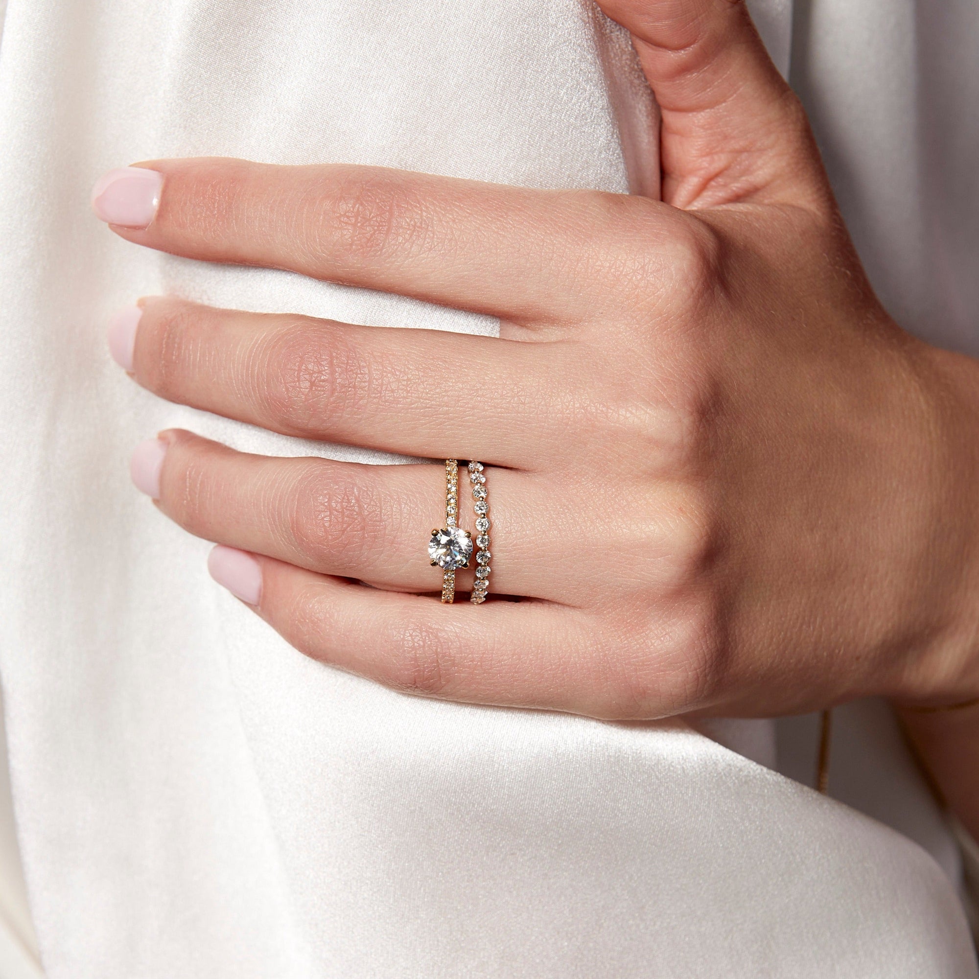 Lifestyle Model Close-up of a woman's hand gracefully resting against a soft white fabric, wearing a beautiful stack of two gold rings on her ring finger: a classic round diamond solitaire ring and a delicate prong-set diamond band.