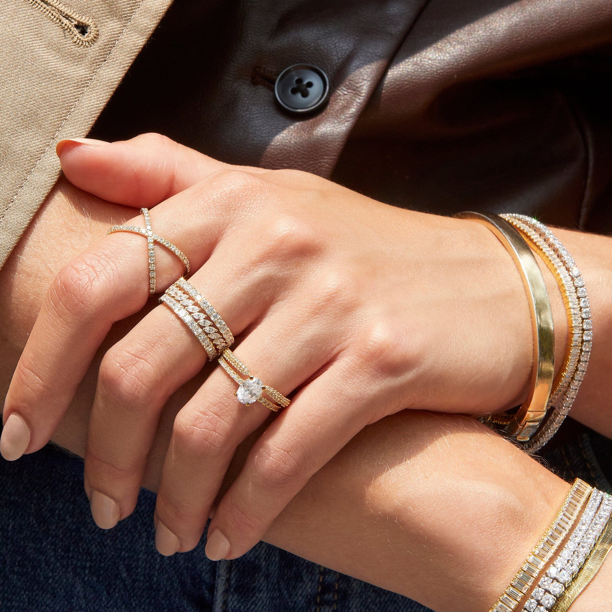Lifestyle Model Close-up of a woman's hands resting against a brown leather garment, wearing multiple stacked gold and diamond rings, including an oval engagement ring and eternity bands, alongside layered gold and diamond bracelets