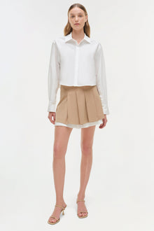 Beige pleated skort with white trim hem, styled with white button-up shirt and tan sandals.