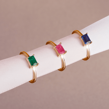 Model Close-up of three gold rings with green, pink, and blue baguette gemstones resting on a white display
