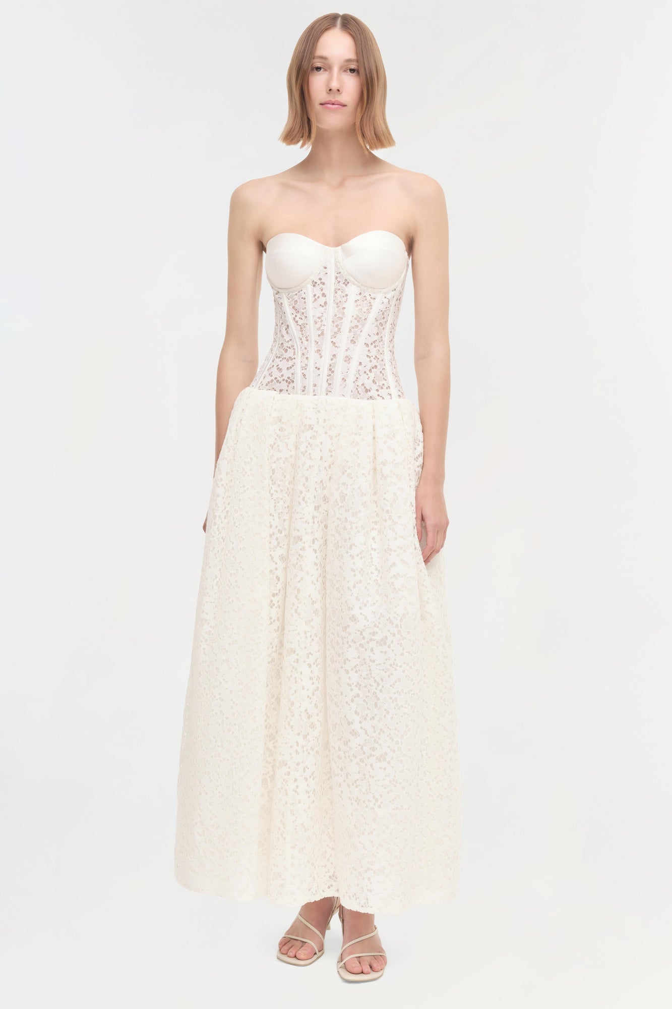 Woman wearing ivory strapless dress with lace corset bodice and full lace skirt, paired with white sandals.