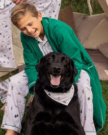A young boy in a green robe and ski-patterned pajamas smiles behind a black Labrador sporting a matching The Great Outdoors bandana from Petite Plume. Both appear happy outdoors, surrounded by pillows and cushions.