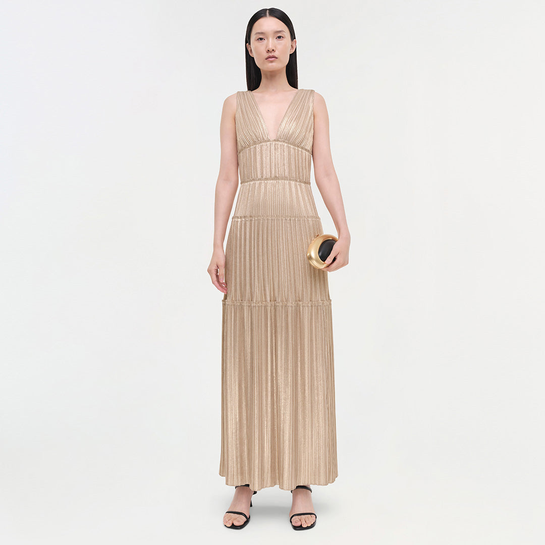 Beige pleated sleeveless gown with deep V-neckline and tiered waistband sections, worn with gold clutch and black sandals.