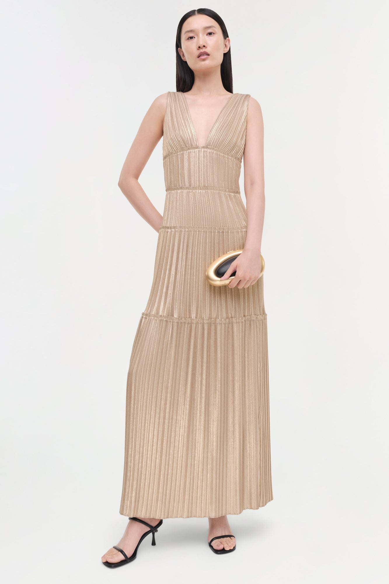 Beige pleated sleeveless gown with deep V-neckline and tiered skirt, worn with gold clutch and black sandals.