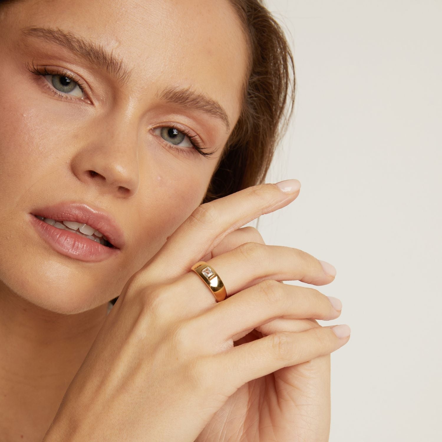 Close-up of a woman's face with a gold ring on her finger against a neutral background