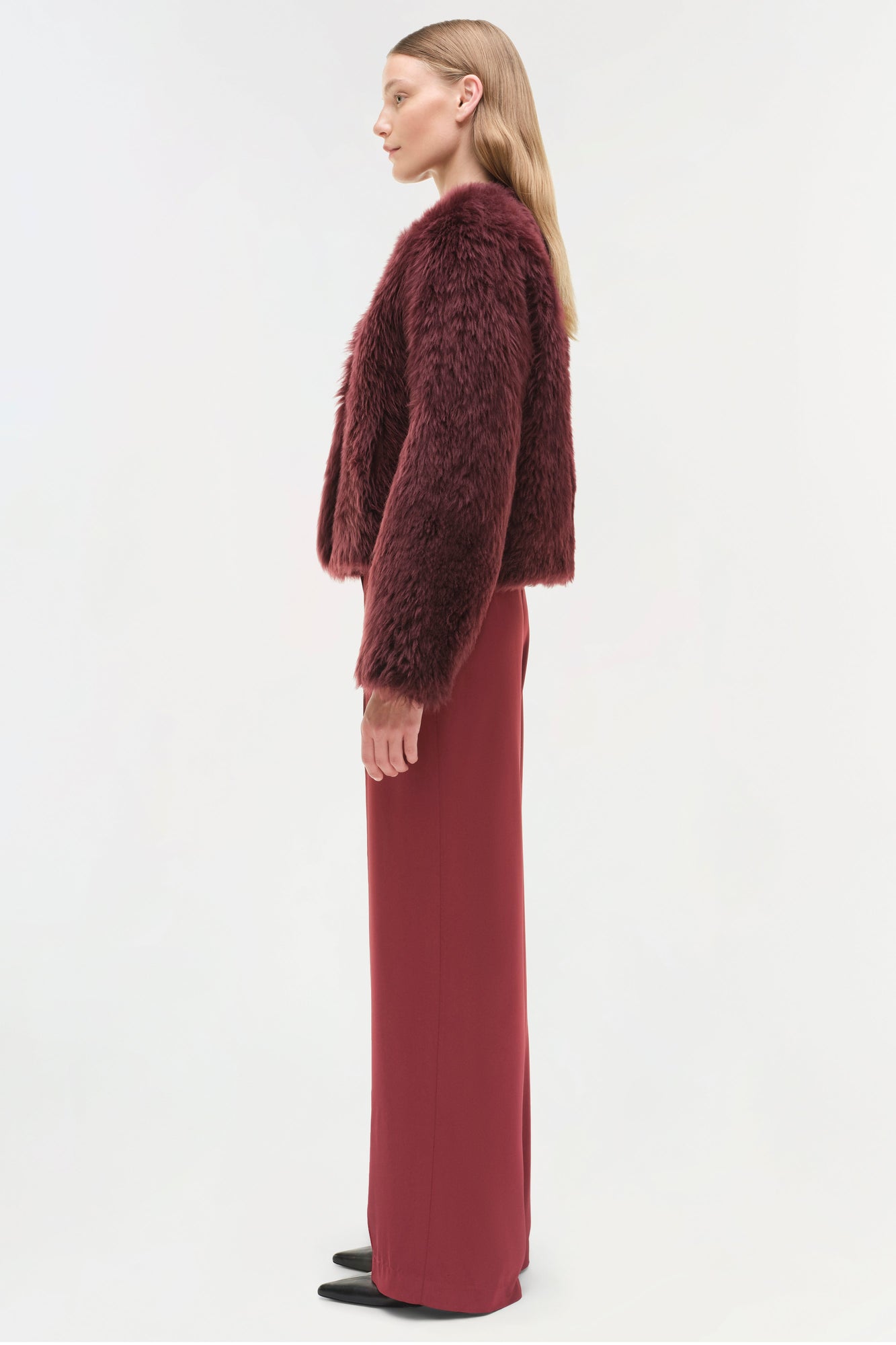 Woman wearing burgundy shearling jacket with cropped fuzzy sleeves over matching long dress, shown from side profile.