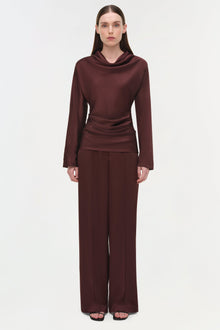 Woman wearing burgundy satin draped long-sleeve top with cowl neckline and ruched waist detail paired with wide-leg pants.