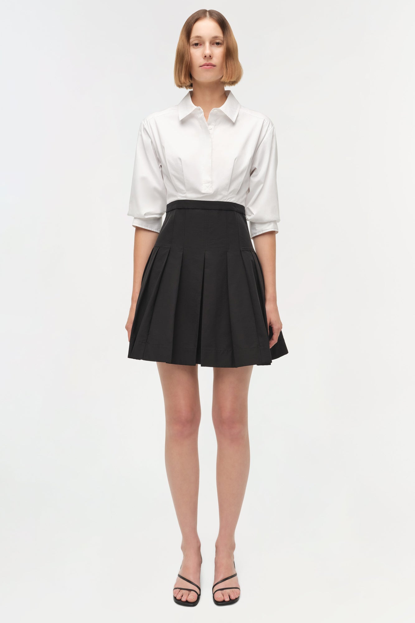 White collared shirt tucked into black pleated mini skirt with three-quarter sleeves.