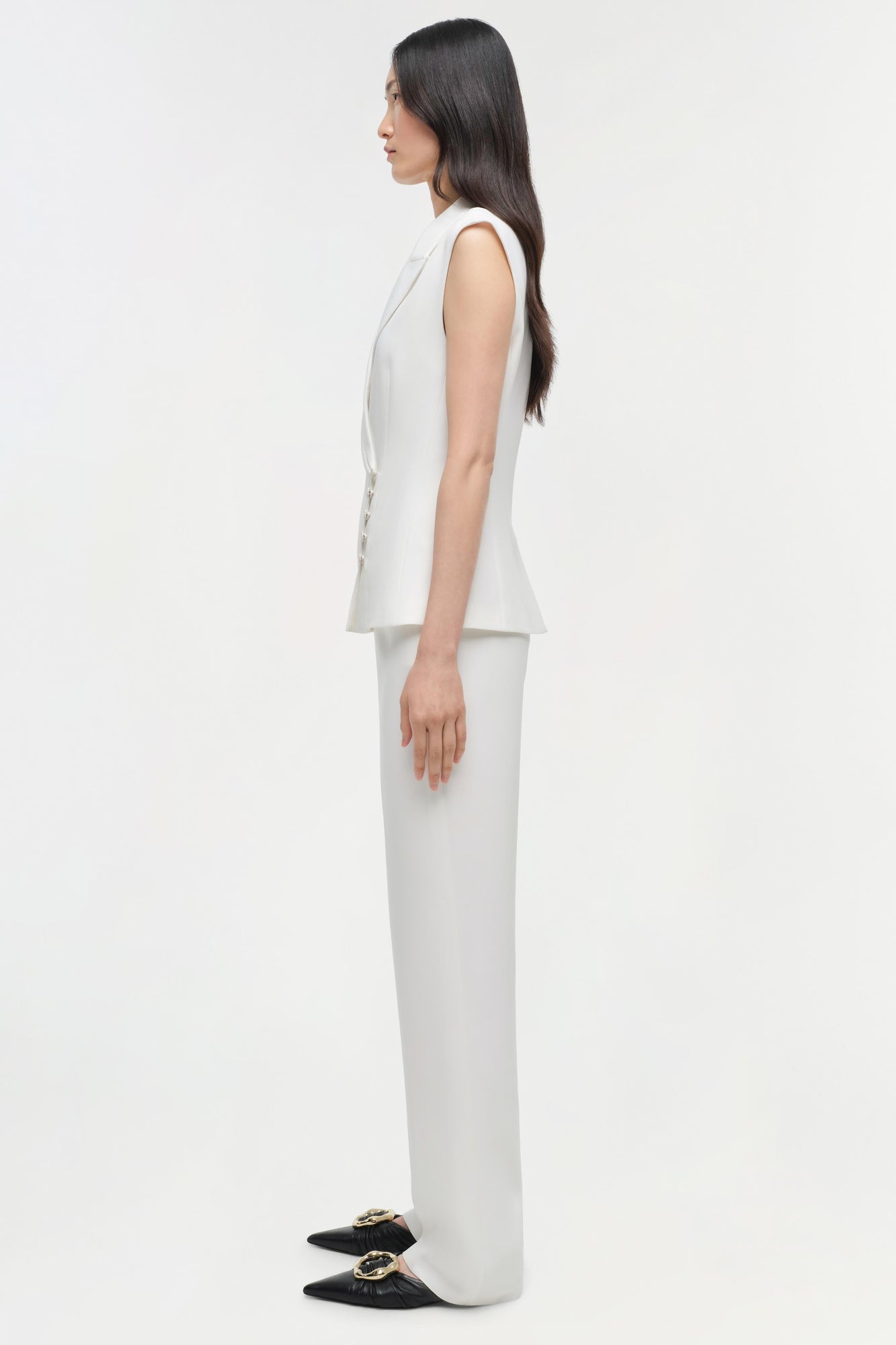Woman wearing cream sleeveless vest and white straight leg trousers with black embellished loafers, side view.