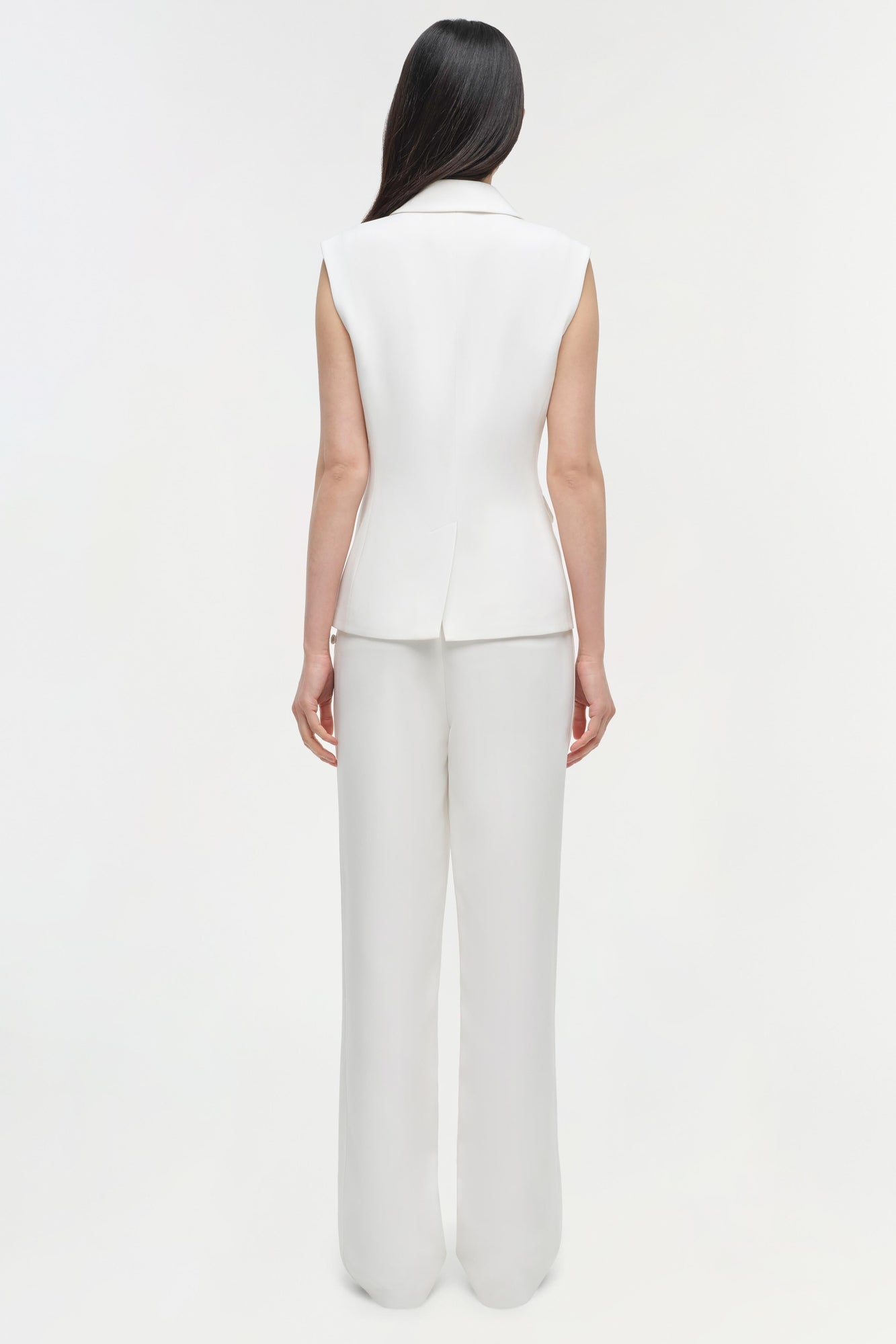 Woman wearing cream sleeveless vest and straight leg trousers in cream, rear view.