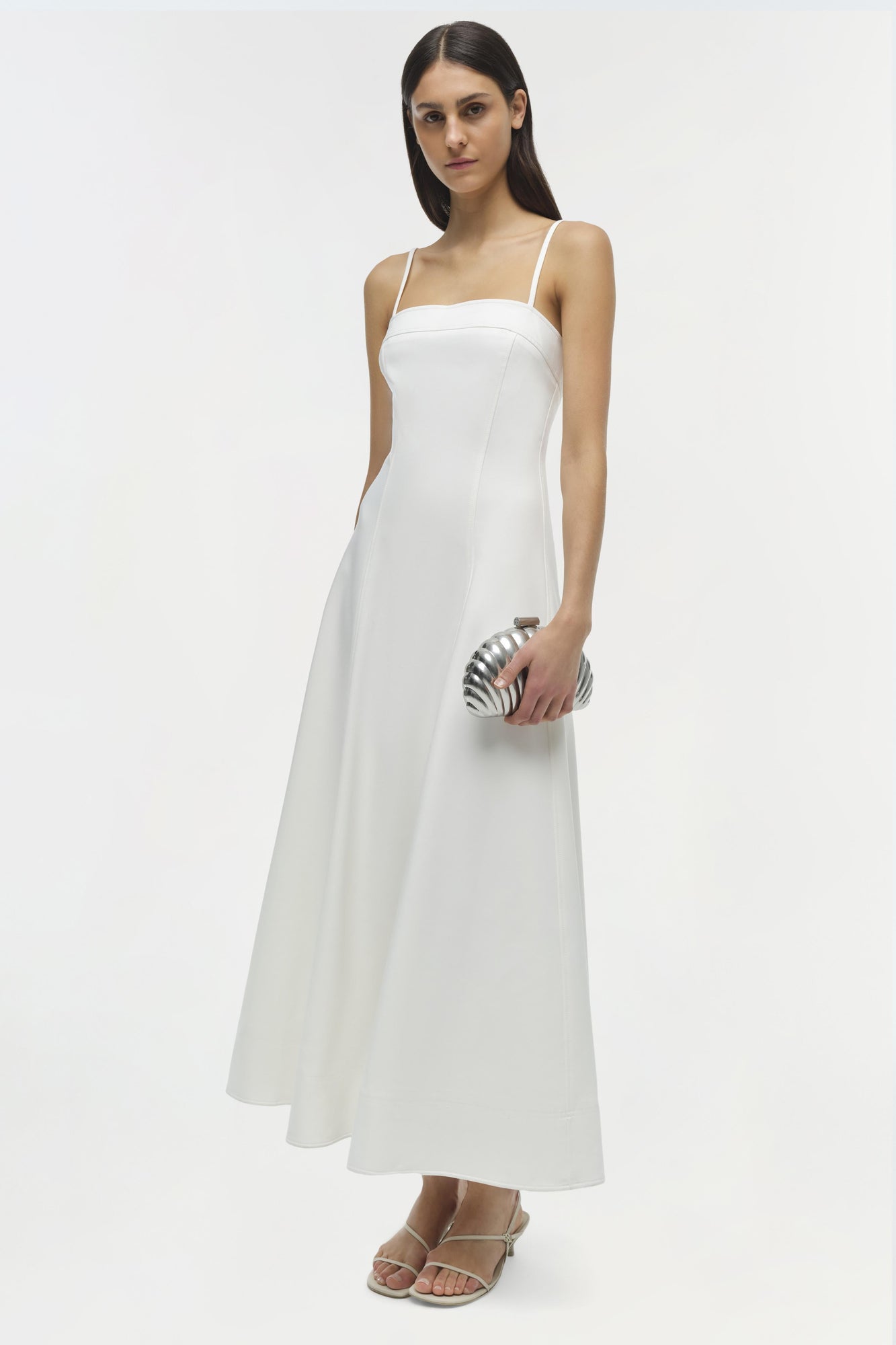 White cotton midi dress with thin straps and fitted bodice on model wearing metallic clutch.
