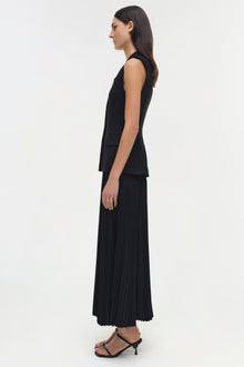 Woman wearing black sleeveless layered dress with pleated skirt and black strappy heels, shown in profile.