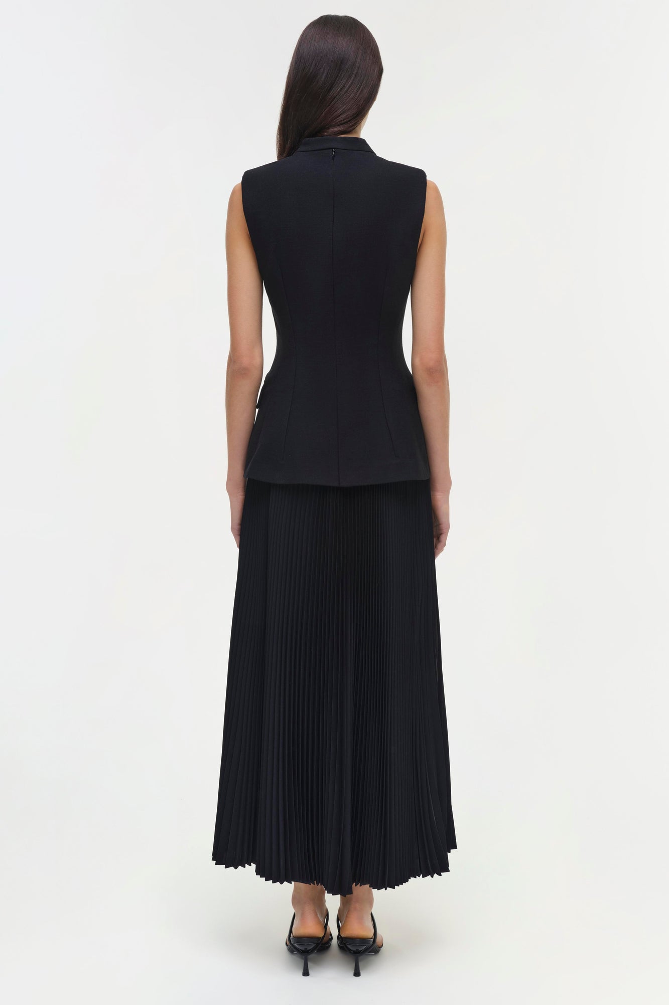 Woman wearing black sleeveless dress with fitted bodice and pleated midi skirt, shown from back view.