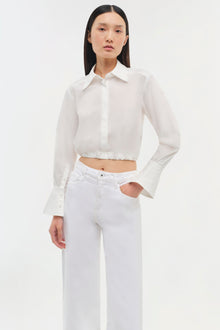 White cropped button-up shirt with long flared sleeves and gathered hem, styled with white jeans.