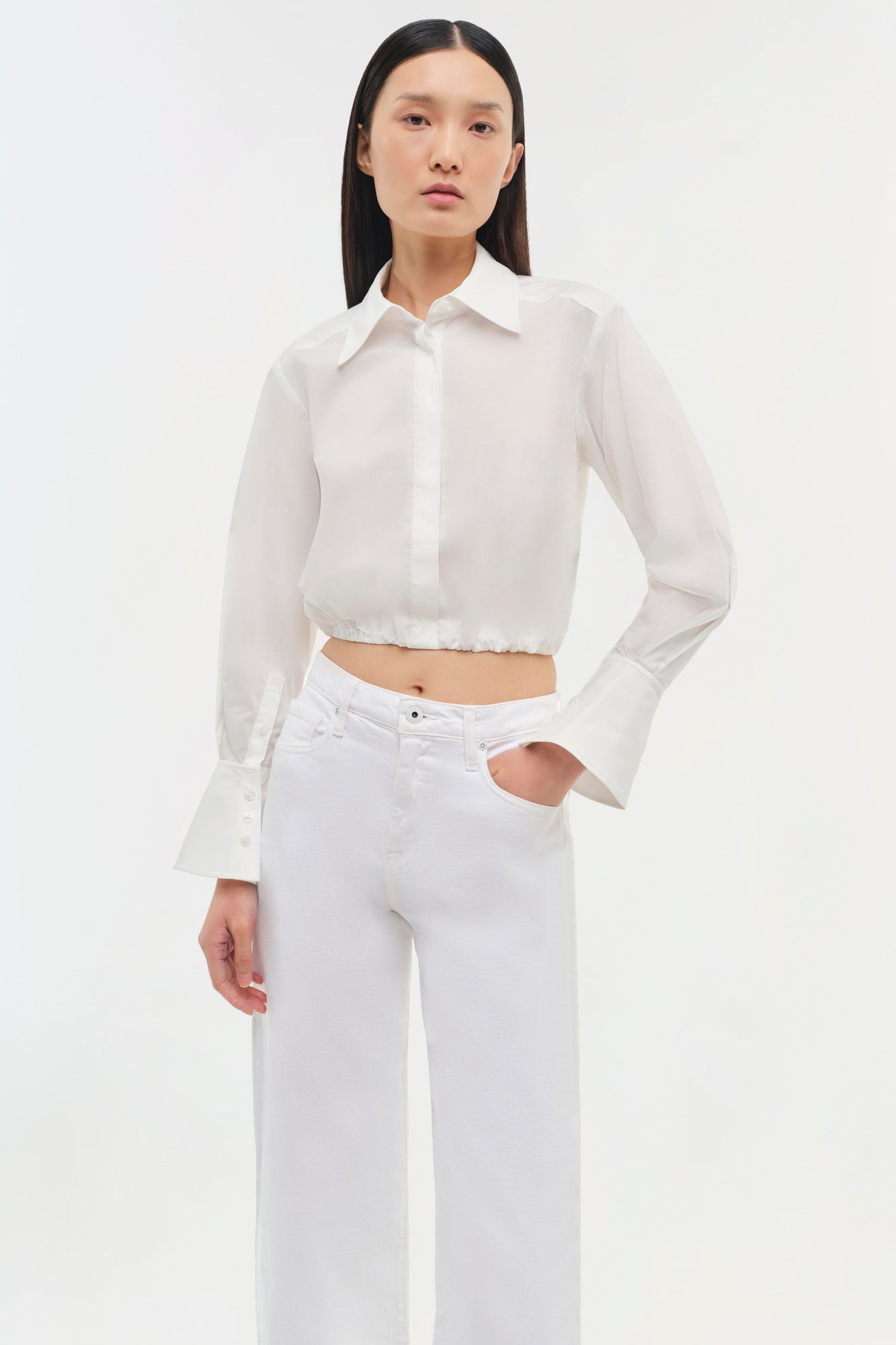 White cropped button-up shirt with long flared sleeves and gathered hem, styled with white jeans.