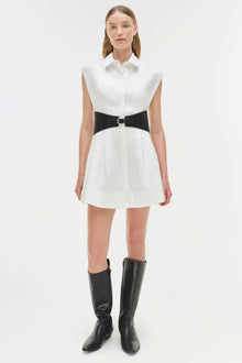 White sleeveless collared cotton mini dress with black wide belt at waist, paired with black knee-high boots.