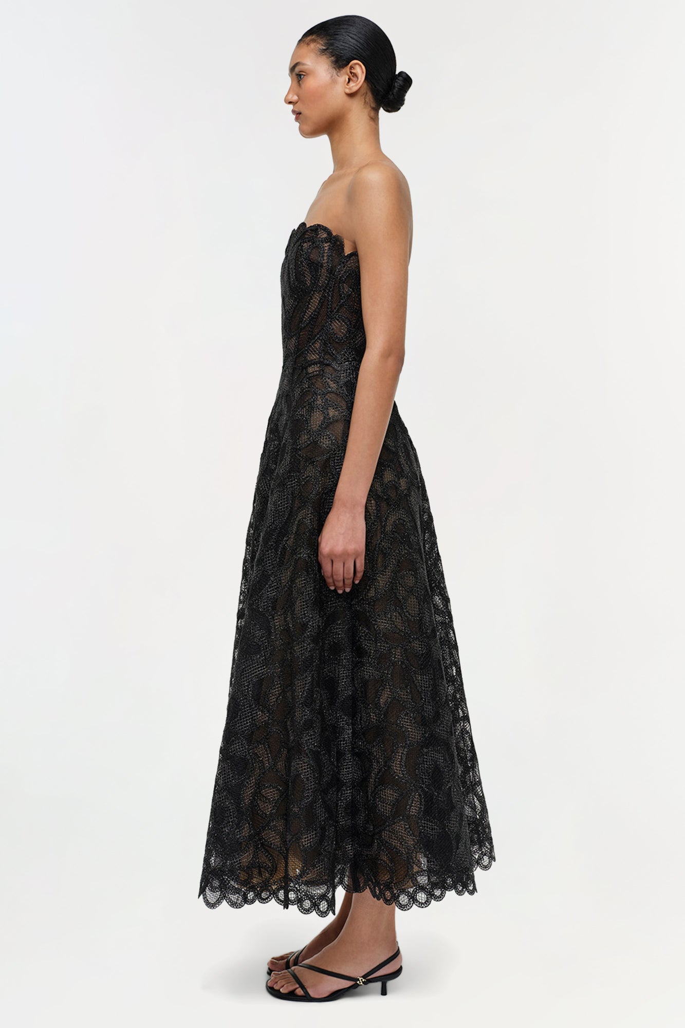 Black strapless lace midi dress with geometric pattern and scalloped hem, shown on model from side profile.