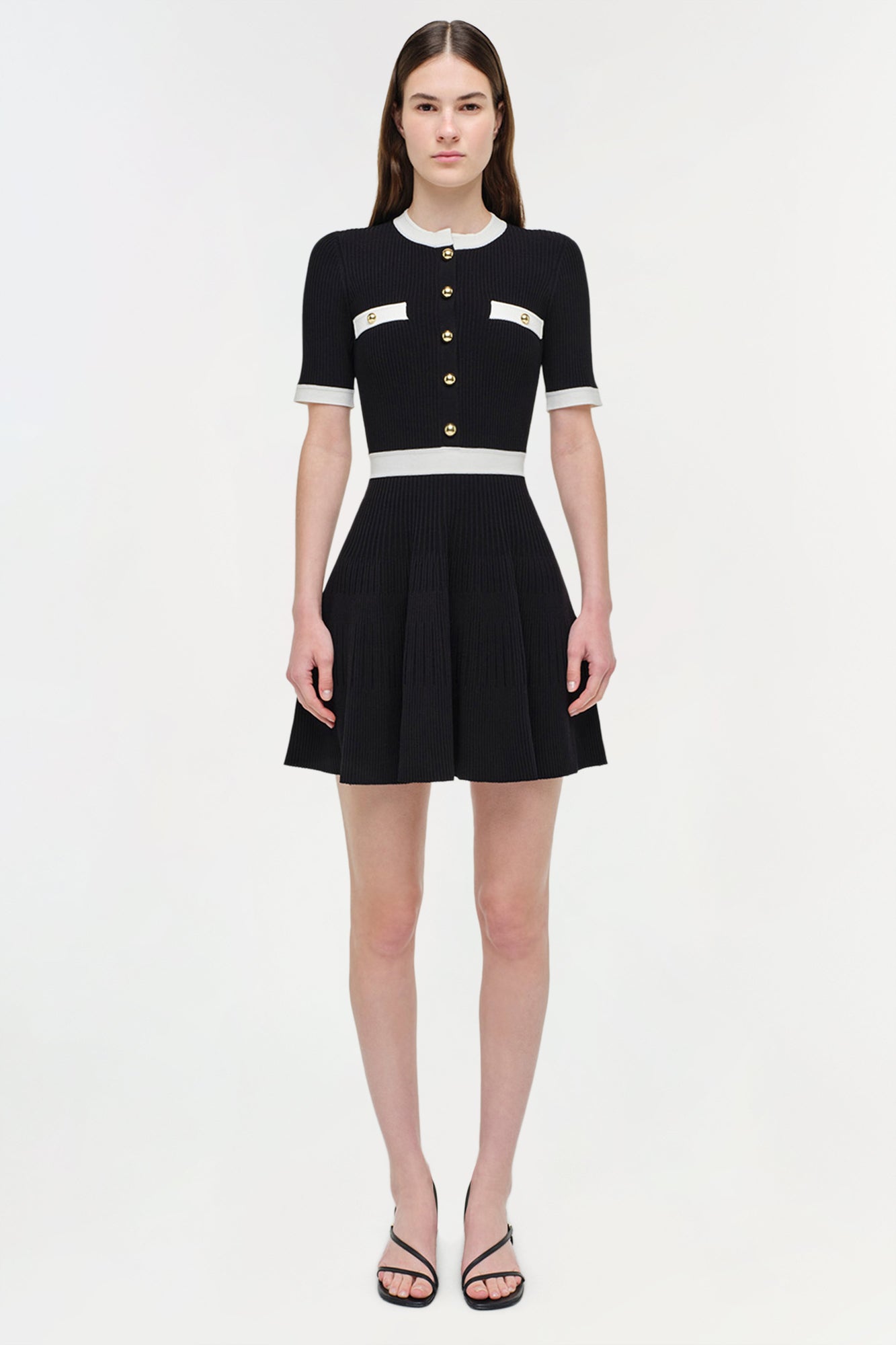 Black ribbed knit mini dress with white trim, gold button front closure, and pleated skirt.