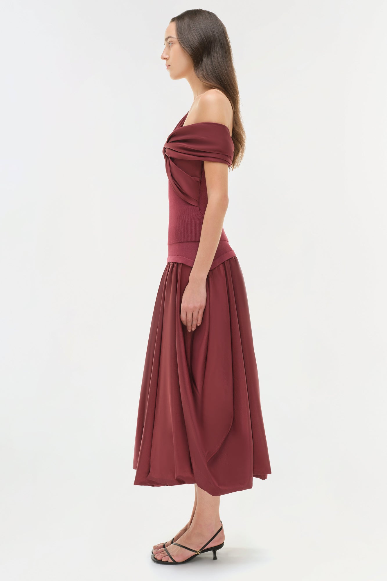Woman wearing burgundy off-shoulder top with draped neckline and full midi skirt, side view.