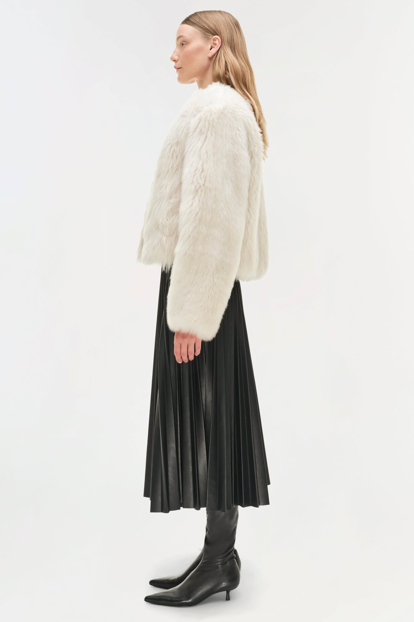 Woman wearing a cream-colored shearling jacket over a black pleated midi skirt and black boots, shown from the side.