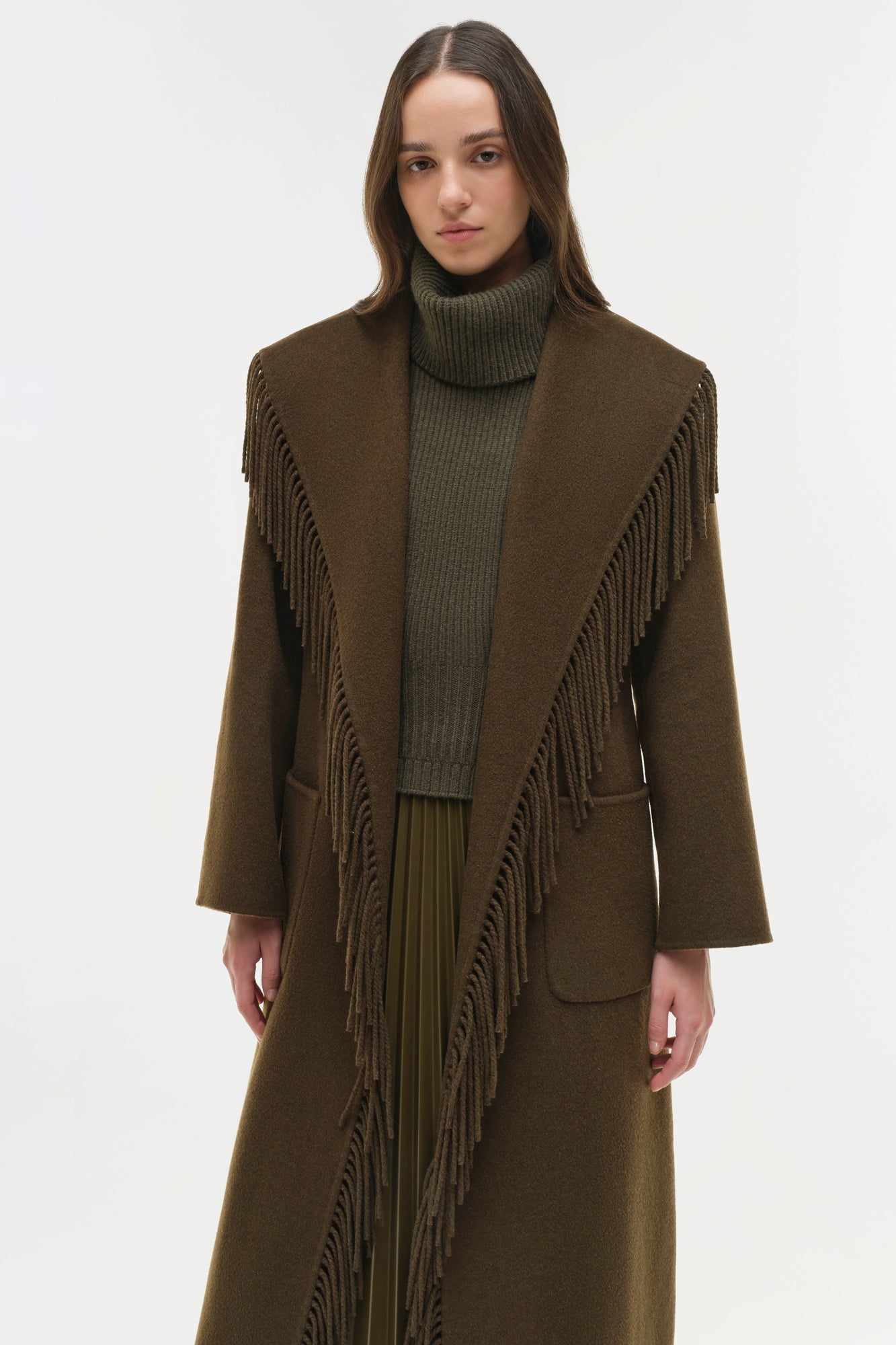 Olive brown wool coat with fringe trim along the front edges and long sleeves, styled over a turtleneck and pleated skirt.