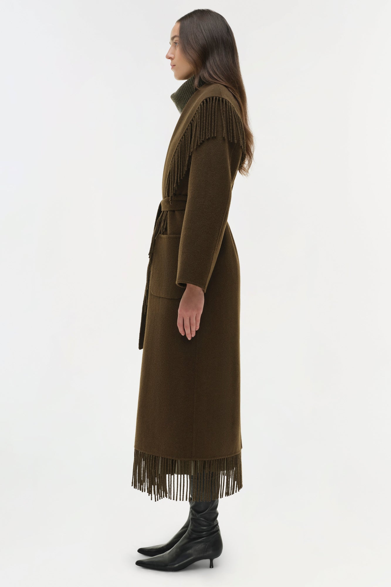 Olive-brown coat with fringe trim at shoulders and hem, belted waist, worn with black boots.