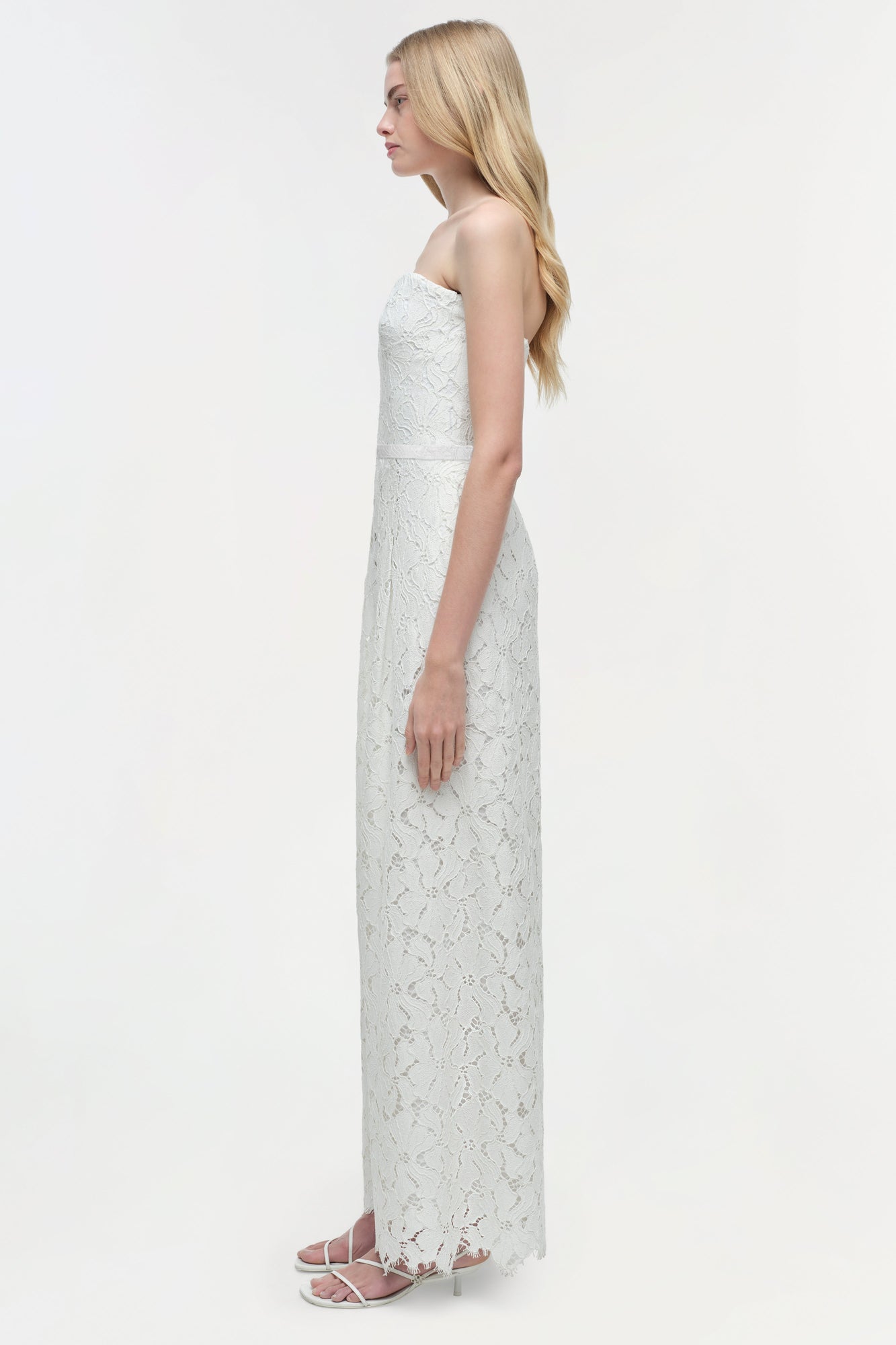 Woman wearing a white lace strapless jumpsuit with wide legs, shown from the side profile.
