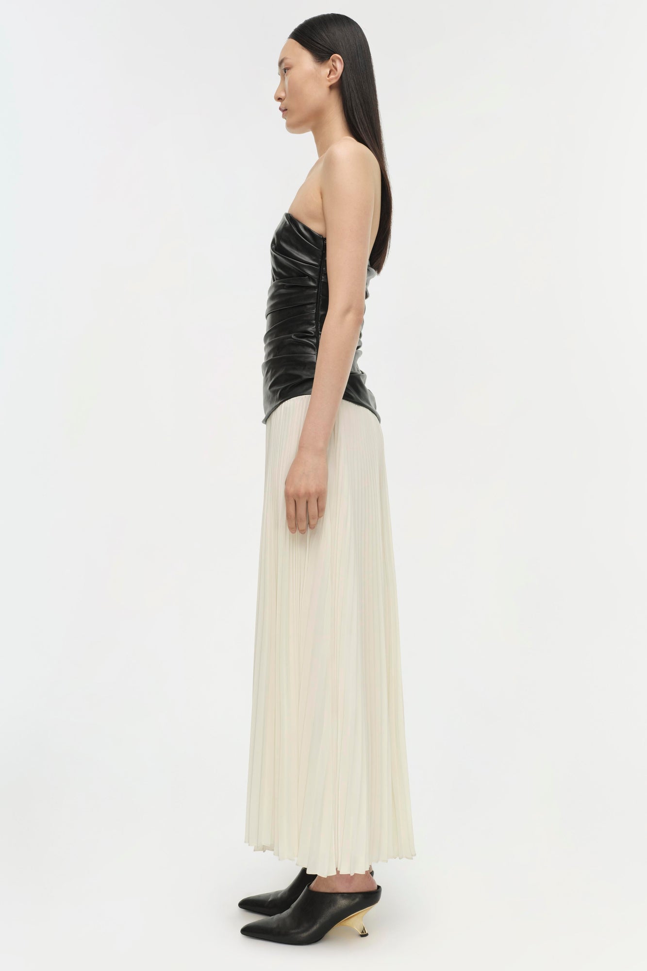 Woman wearing strapless black ruched top with cream pleated skirt and black ankle boots, shown in profile.