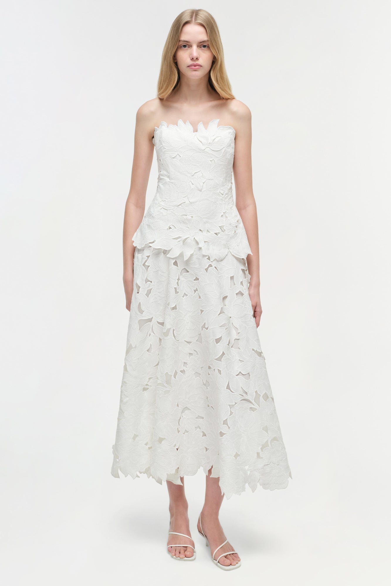 White strapless midi dress with leaf-shaped embroidered details throughout, worn with white strappy sandals.
