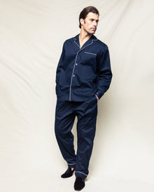 A man wearing Petite Plumes Mens Twill Pajamas in Navy with White Piping stands against a light background, hands in pockets and legs slightly crossed. He looks left while wearing black slippers.