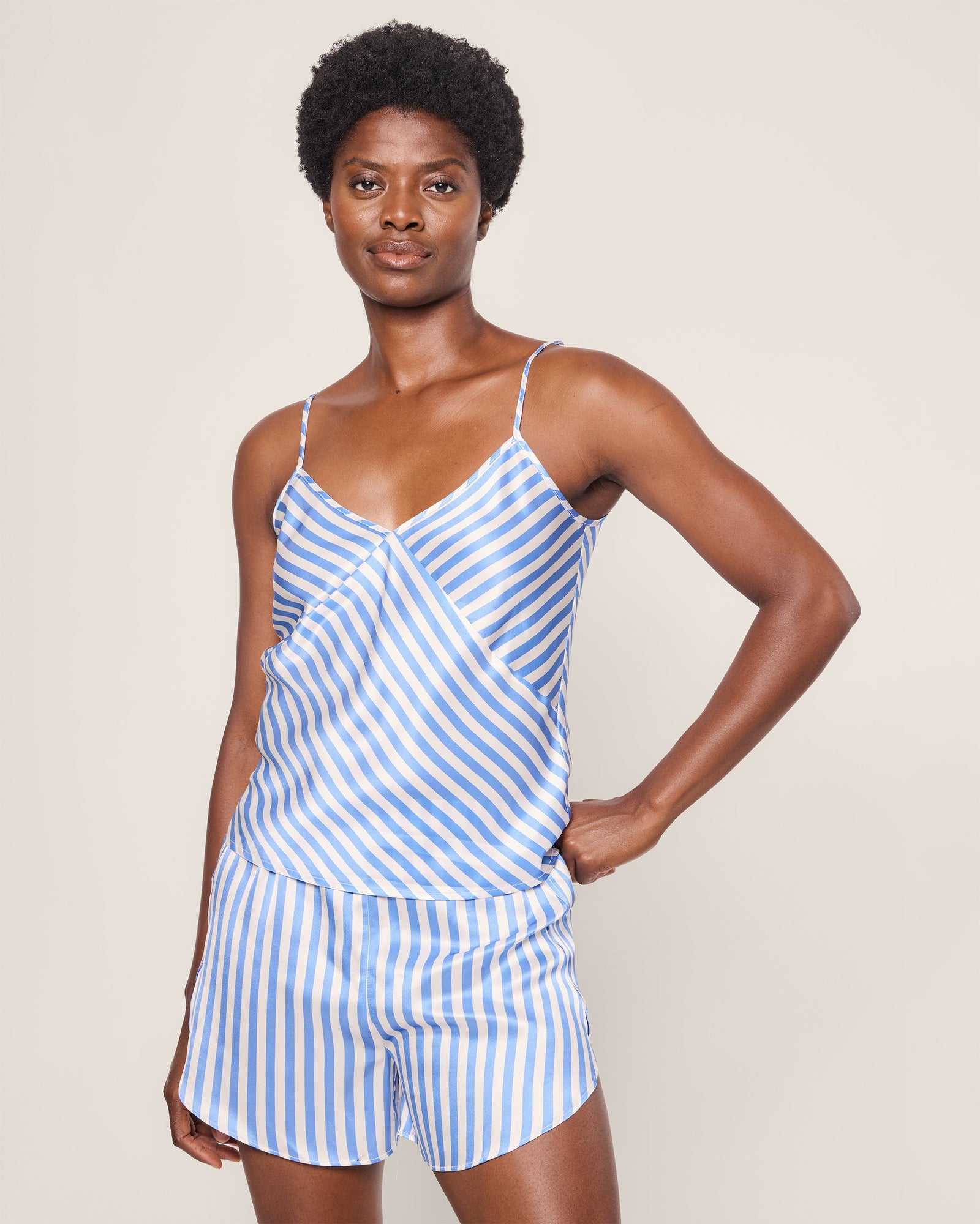 A person with short curly hair stands confidently with one hand on their hip, wearing the Petite Plume Womens Silk Cami Short Set in Azure Stripe. The plain, neutral background highlights the elegance of their luxury sleepwear.