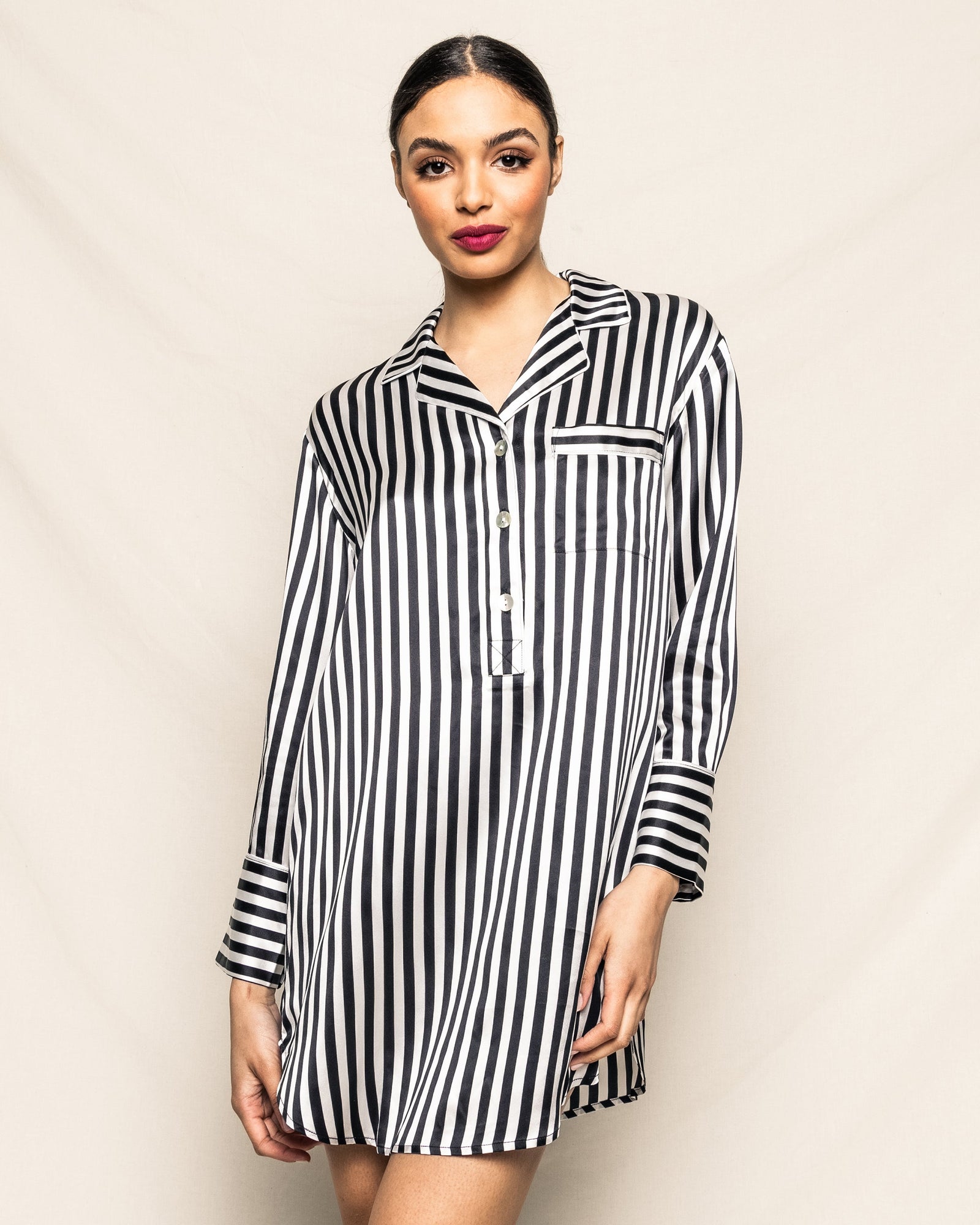 A woman stands against a light beige background, looking at the camera with her hair tied back and bold red lipstick, wearing the Petite Plume Womens Silk Nightshirt in Black and White Bengal Stripe.