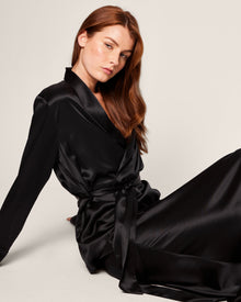 A woman with long auburn hair is reclining against a light backdrop, wearing a black Womens Silk Short Robe from Petite Plume. The robe comes with a matching belt, highlighting her relaxed and elegant demeanor—ideal for luxurious sleepwear.