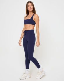 Side view of the LSPACE Uppercut Bra in midnight blue on a model against a white background, showing the square-neck sports bra profile with the lace-up side waistband detail of the matching midnight blue active pants, white sneakers and socks.