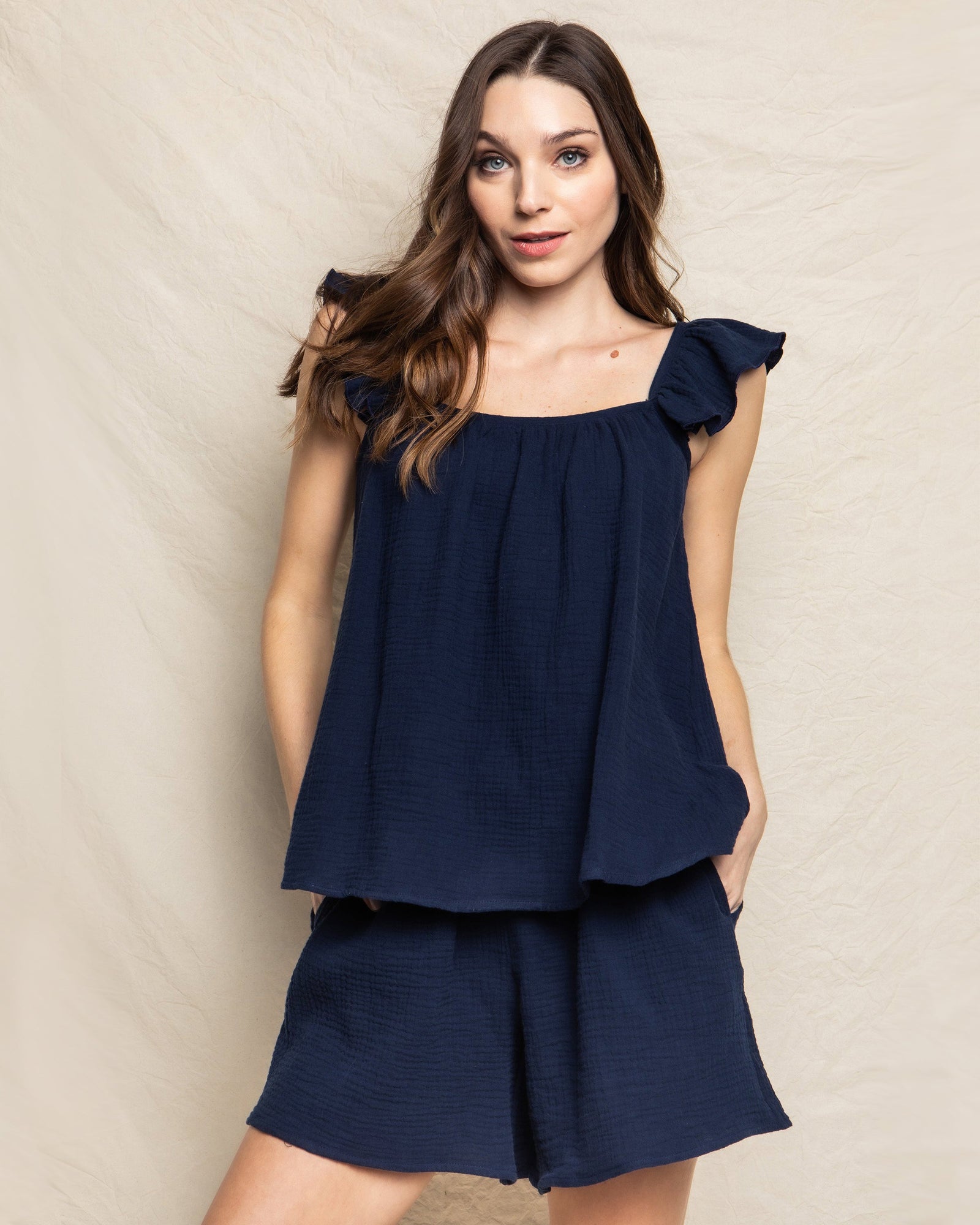 A woman with long hair wears a sleeveless, loose-fitting Womens Gauze Celeste Top in Navy by Petite Plume and matching shorts against a light, textured backdrop. Her hands in pockets, she gazes at the camera with a slight smile, embodying elegant feminine silhouettes.