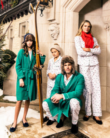 Four people in luxurious sleepwear pose on a snowy porch. One has wooden skis, another wears a red scarf, and all sport fur hats. The ornate building features stone details and a lantern above, enhancing the coziness of their Petite Plume Womens Flannel Pajama Set in Apres Ski.