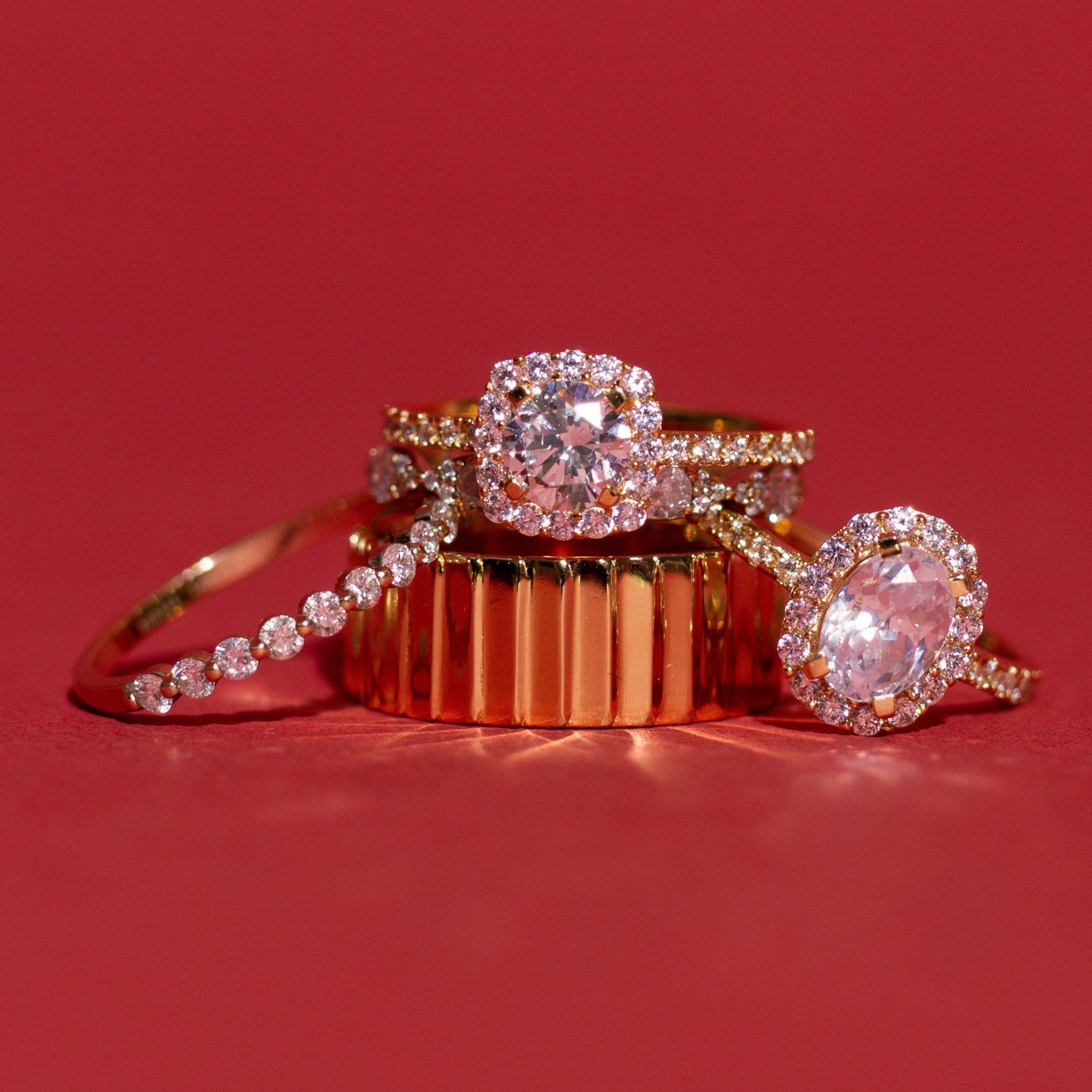 Flatlay Model Close-up of four gold and diamond rings arranged against a deep red background. The collection features a delicate single-row diamond band, a thick ribbed gold band, a cushion-cut halo diamond ring, and an oval-cut halo diamond ring.