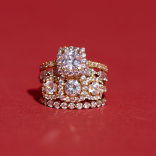 Flatlay Model Close-up of a dazzling stack of three gold and diamond rings resting against a deep red background. The stack features a large cushion-cut halo diamond ring on top, a three-stone round diamond halo ring in the middle, and a thick prong-set diamond band at the bottom.