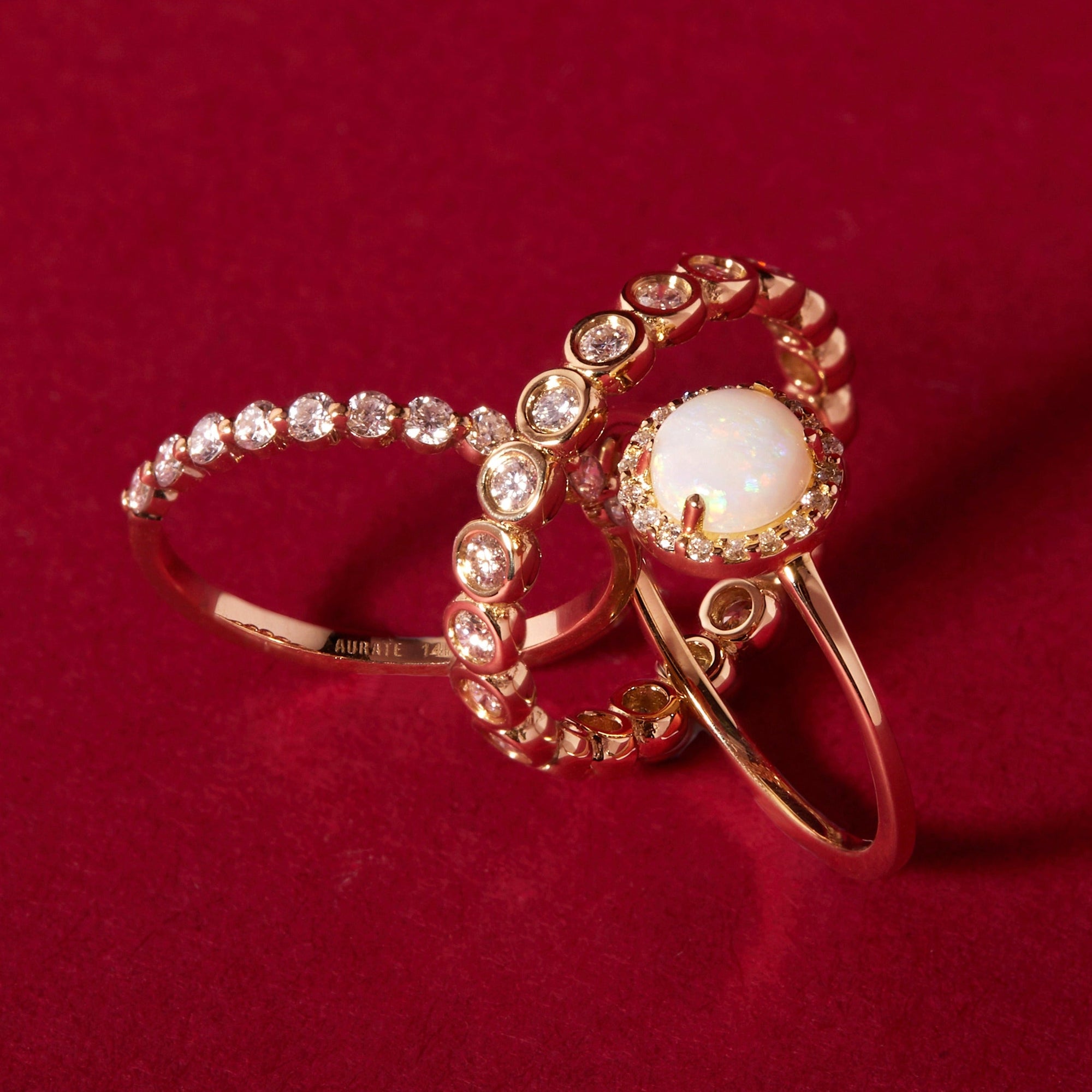 Flatlay Model Close-up of three intertwined rose gold rings resting against a deep red background. The rings include a delicate prong-set diamond band, a bezel-set diamond band, and a ring featuring a prominent round opal surrounded by a diamond halo.