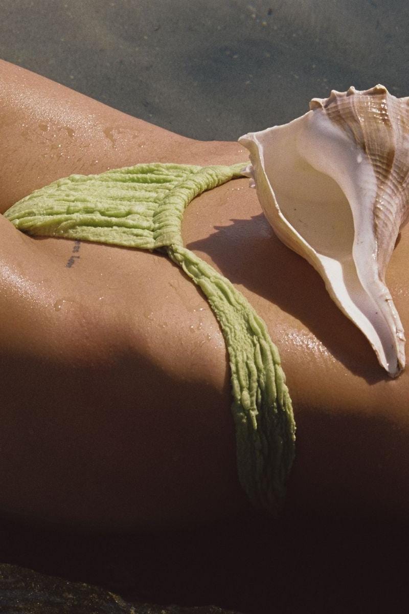 Green textured bikini bottom with tassel tie detail on model's hip at beach.