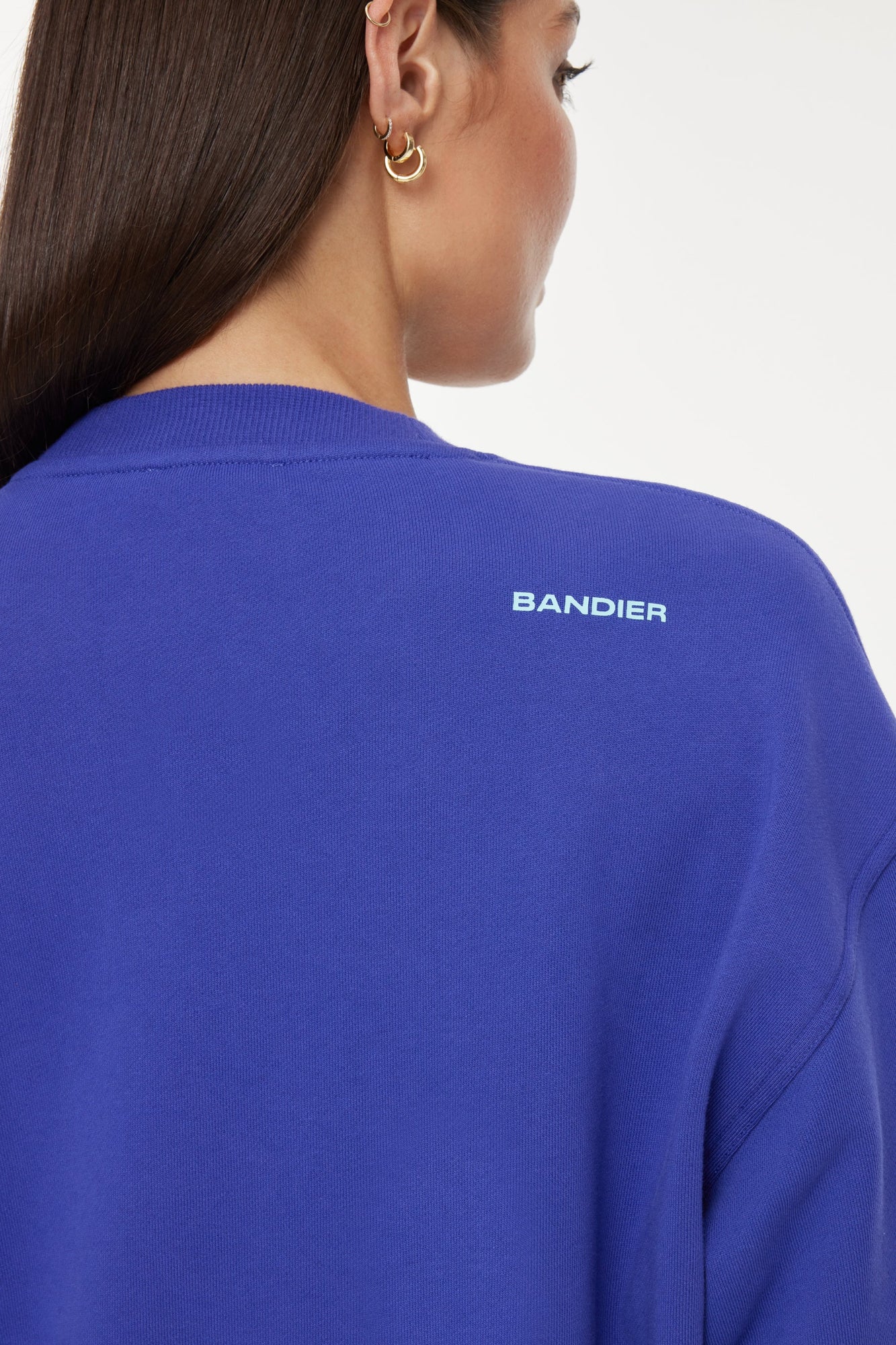 Model wears a royal blue crewneck sweatshirt with the lettering "Soleil Sport" across the chest with wavey lines below and "Bandier" in the bottom right corner. 