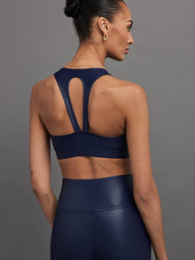 Model wears a navy, glossy, deep scoop-neck sports bra with a sleek racerback and a shiny, liquid-like finish.
