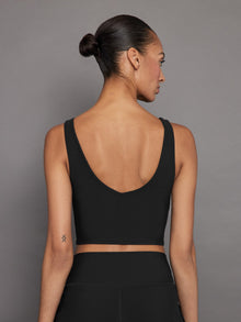 Model wears a black, longline sports bra with a deep V-neck front and an open V-shaped back, made from sleek, smooth fabric.
